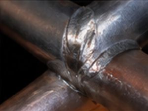Close-up of a welded cross joint on metal pipes with shiny weld bead and heat discoloration around the seam.