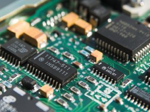 Close-up of a green printed circuit board with black integrated circuit chips and orange capacitors, detailing dense electronic components in a motherboard-like layout.